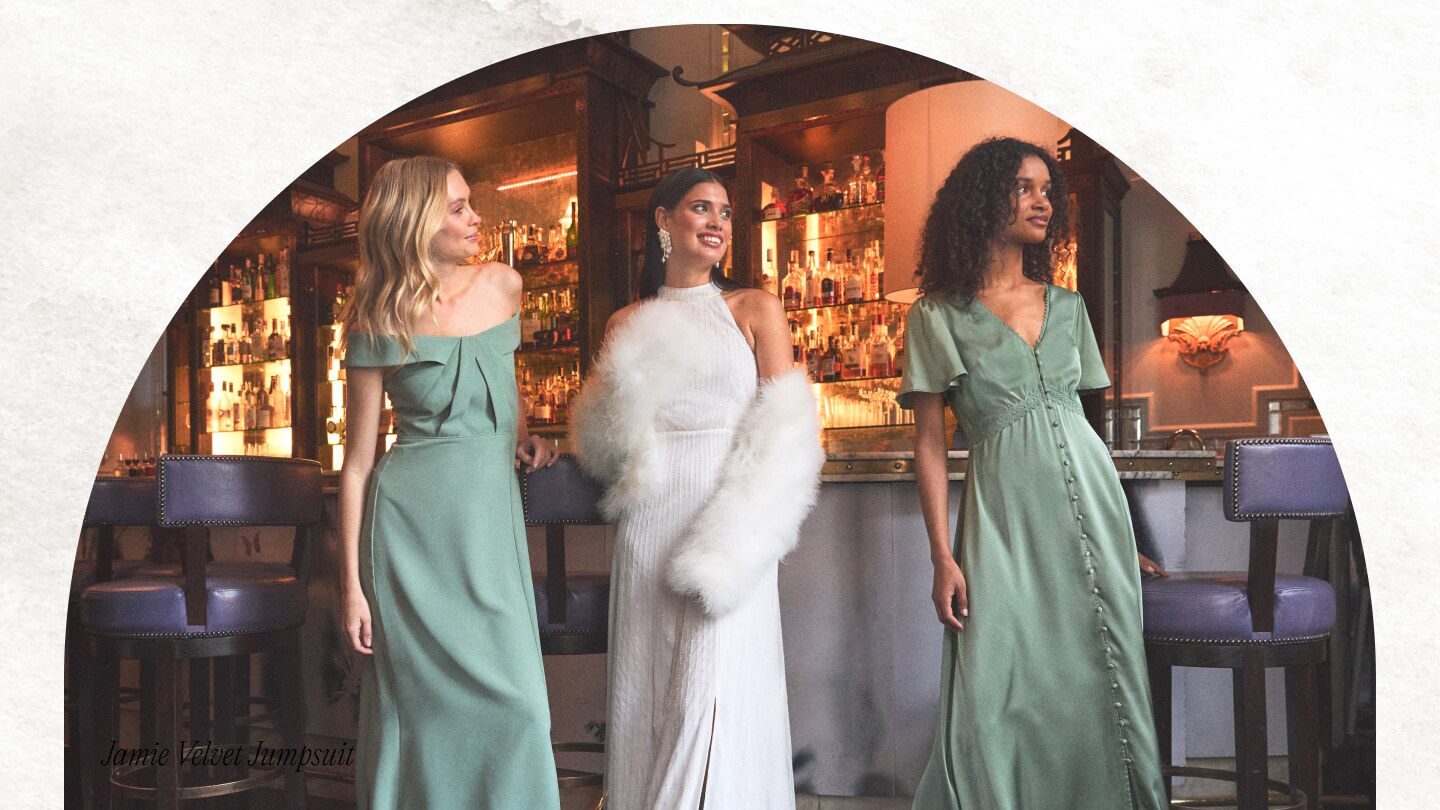 Bride wears white halterneck wedding dress with white furry wrap in an ambient bar with her two bridesmaids in sage green dresses